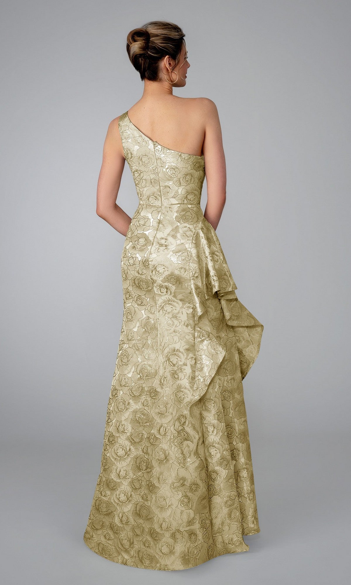 A woman faces away from a plain gray background, wearing the Stella Couture 26315 long formal dress in champagne, featuring a gold one-shoulder design and textured floral brocade for an elegant evening look.